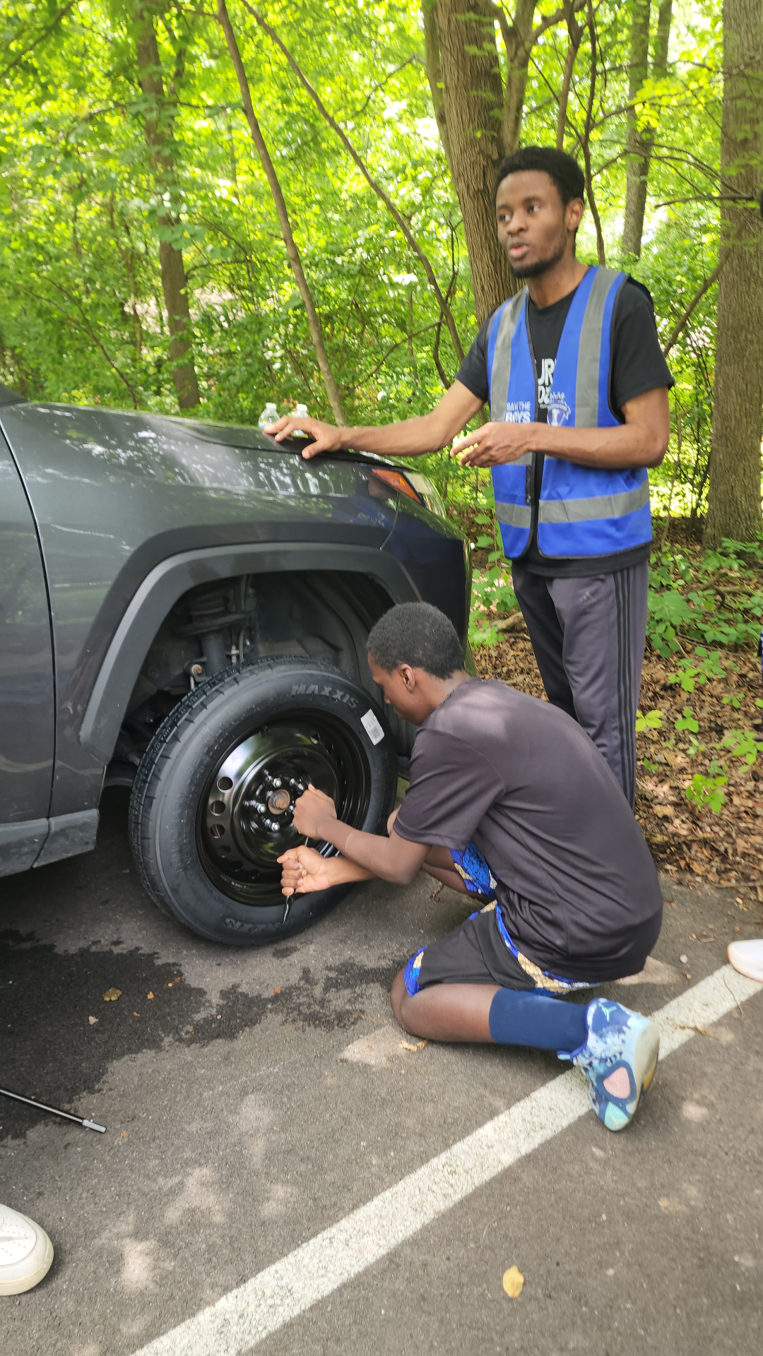 A BLA volunteer in a Save the Boys vest coaches a young man through changing a tire at camp.