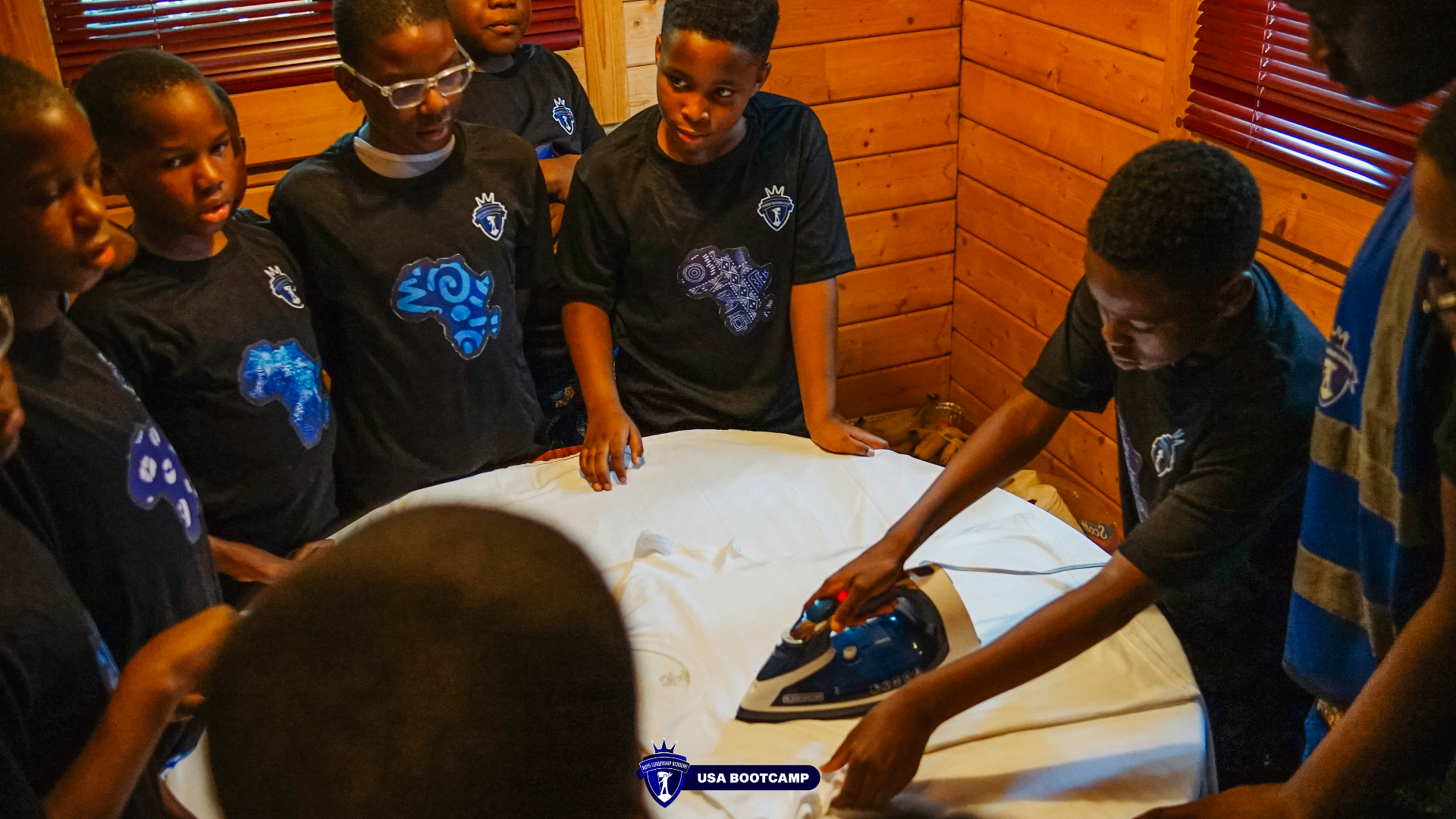 Boys learning to iron a shirt inside a cabin during boot camp.